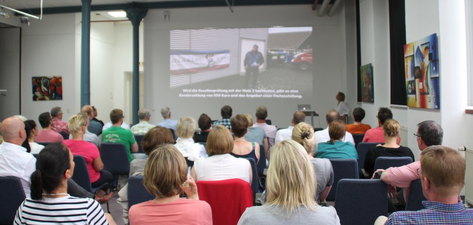 Ein Publikum sitzt auf Stühlen vor einer Bühne mit Leinwand.