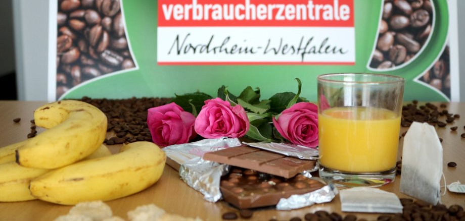 Bananen, Kaffeebohnen, Rosen, Schokolade, Teebeutel, Kekse und ein Glas Orangensaft auf einem Tisch. Im Hintergrund das Logo der Verbraucherzentrale Nordrhein-Westfalen.