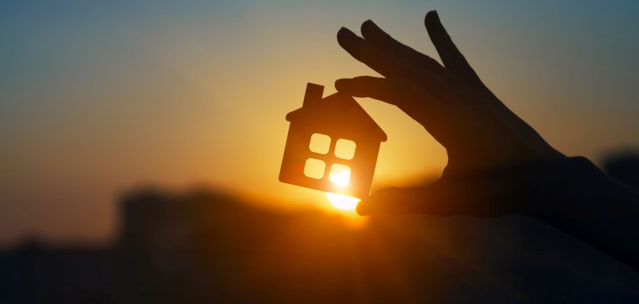 Eine Hand hält ein kleines Haus aus Holz vor den Sonnenuntergang.