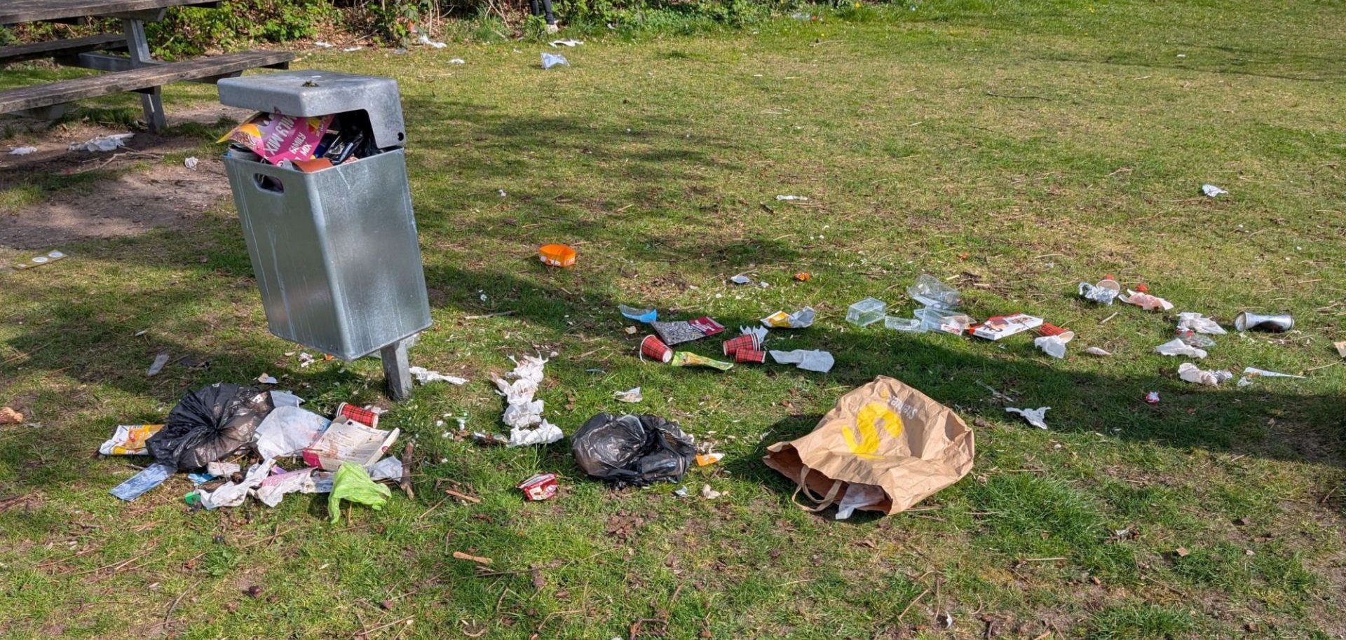 Überfüllter Mülleimer mit herumliegendem Abfall auf einem Spielplatz.
