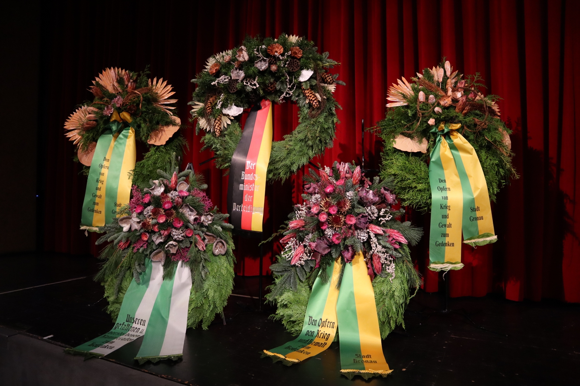 Kränze mit Trockenblumen und Gedenkbändern vor einem roten Vorhang.