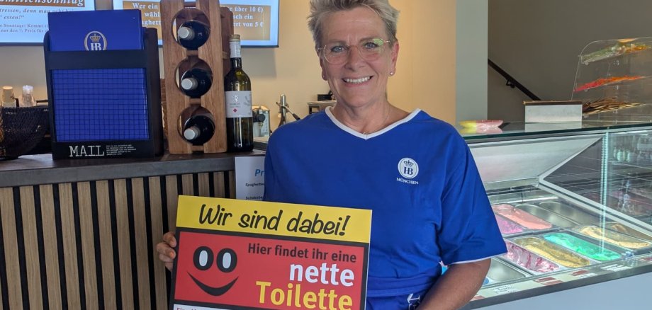 Das Bistro Gustav ist ein neuer Partner bei der Netten Toilette in Gronau. Eine Frau des Bistro Gustav hält ein Schild mit der Aufschrift "Wir sind dabei! Hier findet ihr eine nette Toilette."
