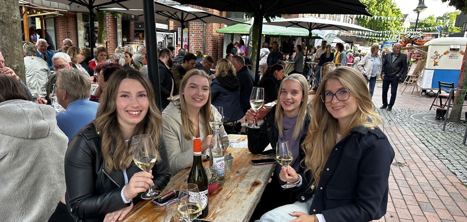 Vier Personen sitzen an einem Tisch und trinken Wein. Sie heben ihre Gläser und lächeln in die Kamera.
