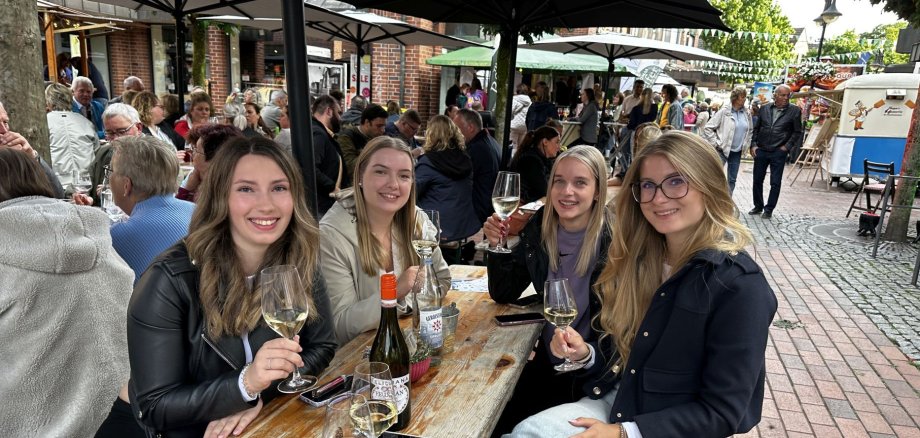 Vier Personen sitzen an einem Tisch und trinken Wein. Sie heben ihre Gläser und lächeln in die Kamera.