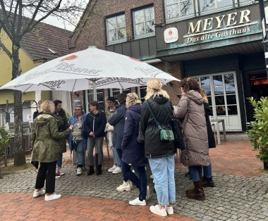 Bei der 'Kneipen, Knifte & ein Korn-Tour' durch Epe wird unter anderem das Gasthaus Meyer besucht. Eine Gruppe von Menschen steht vor dem alten Gasthaus Meyer in Epe. Die Tourführerin erzählt den Teilnehmenden Anekdoten während der Kneipe, Knifte und ein Korn - Tour.